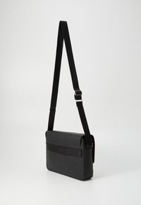 Sac messager en cuir synthétique noir de forme horizontale, avec une sangle noire ajustable et un rabat supérieur comportant une poche avant.