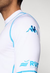 Camiseta deportiva blanca con detalles azules, que presenta un logo bordado en la manga y costuras contrastantes a lo largo de las uniones.