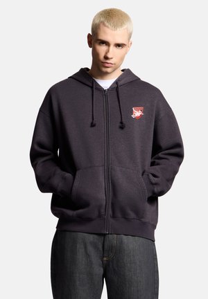 Młody mężczyzna o krótkich blond włosach nosi ciemną bluzę z kapturem i zamkiem, z czerwono-białym emblematem, ręce w kieszeniach, stoi na jednolitym jasnym tle.