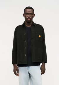 Jachetă neagră din catifea cu guler larg, închidere frontală cu nasturi, două buzunare și un buzunar la piept cu o etichetă galbenă. Poartă-o peste o cămașă neagră.