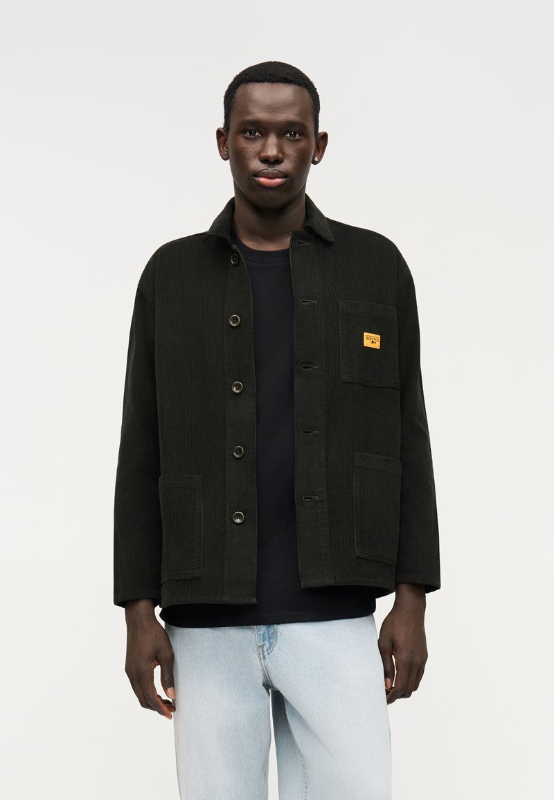 Μαύρο σακάκι από κορδούρα με γιακά άνοιγμα, κλείσιμο με κουμπιά μπροστά, δύο τσέπες και μια τσέπη στο στήθος με κίτρινη ετικέτα. Φορώντας με μαύρο πουκάμισο.