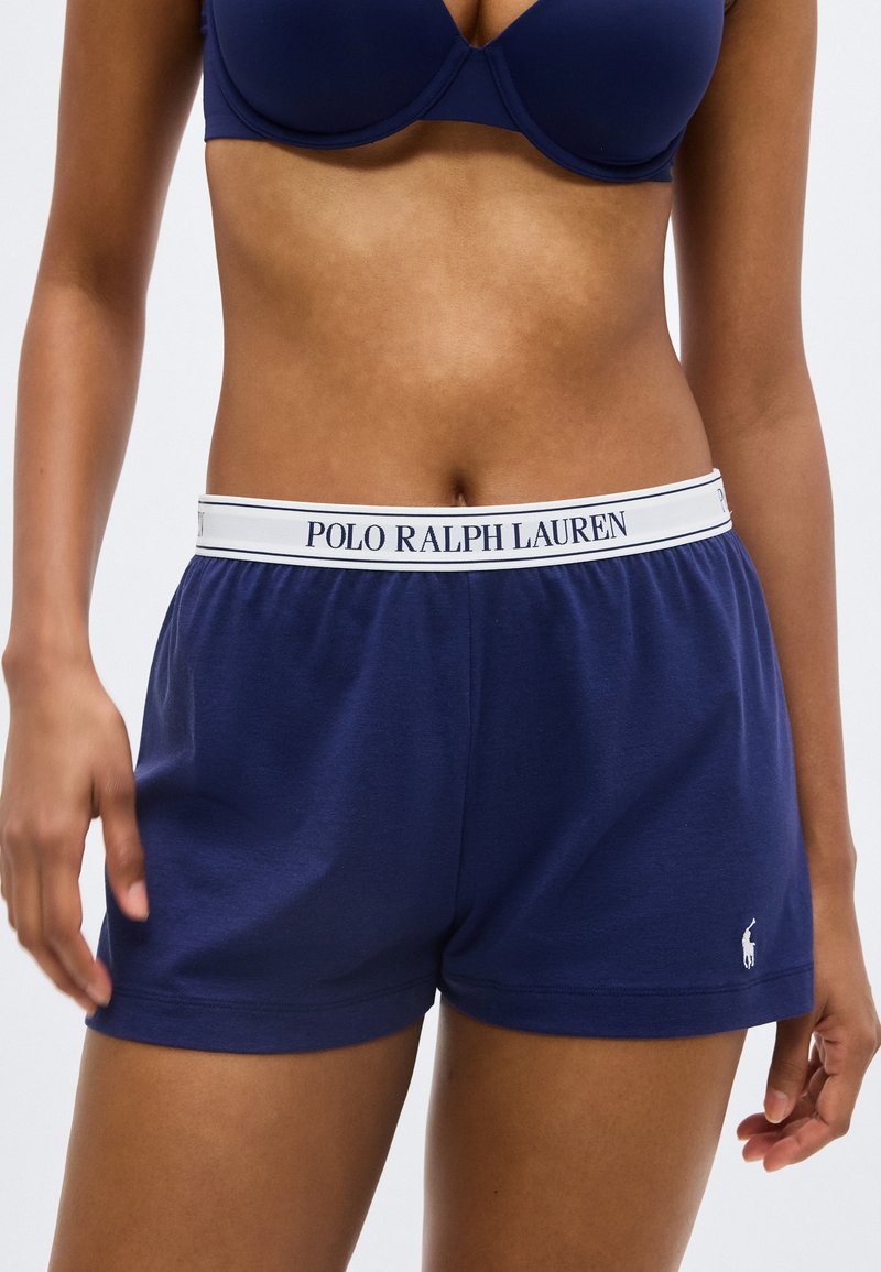 Γυναίκα που φοράει σορτς Polo Ralph Lauren σε ναυτικό μπλε με λευκή ελαστική μέση και σουτιέν σε ναυτικό μπλε, στέκεται μπροστά σε απλό φόντο.