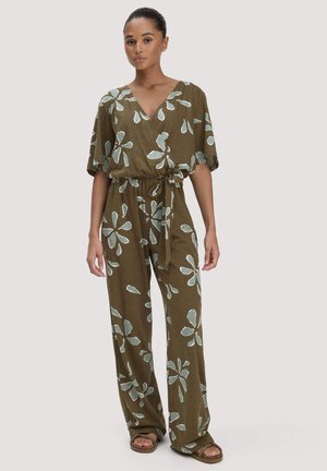 Frau trägt einen olivgrünen Overall mit hellblauen Blumenmustern, kurzen Ärmeln, V-Ausschnitt, gebundenem Taillengürtel und Sandalen, steht vor einem schlichten Hintergrund.