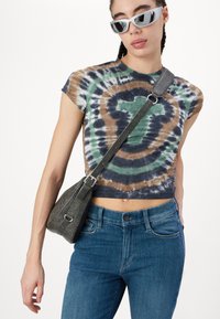 Majica s vezom u stilovima tye-dye u zelenoj, smeđoj i mornarskoj plavoj boji sa kružnim uzorcima, uparena s plavim farmerkama i tamnosivom torbom na preklop. Okvrti bijeli sunčani naočale.