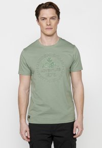 Zaļš īssvārku T-krekls, izgatavots no kokvilnas, ar fakturētu grafisko apdruku, kurā ir uzraksti "KRSH" un "ADVENTURE 1976" kontrastējošās krāsās.