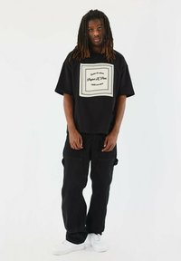 Camiseta negra de gran tamaño con texto gráfico blanco centrado, combinada con pantalones cargo negros y zapatillas blancas. Fondo liso.