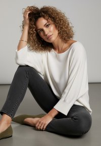 Weißer Pullover mit langen Ärmeln und weitem Ausschnitt, kombiniert mit dunkelgrauer Jeans. Olivgrüne Ballerinas, strukturiertes Haar, minimaler Schmuck. Neutraler Hintergrund.