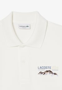 Vit klassisk passform polo tröja med två knappar, Lacoste-märket, och broderad bergs- och Lacoste-logga på bröstet.