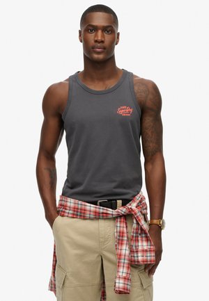 Débardeur en coton gris avec un logo "SuperDry" rouge, porté avec un short cargo et une chemise à carreaux rouges nouée à la taille.