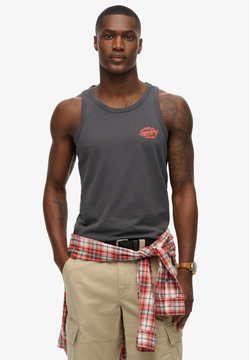 Top de algodón gris sin mangas con un logo rojo de "SuperDry", llevado con pantalones cortos cargo y una camisa de cuadros roja atada a la cintura.