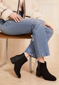Persona vistiendo jeans de un azul claro y botas de ante negras hasta el tobillo, sentada con las piernas cruzadas en una silla marrón, con una chaqueta de borreguito beige.