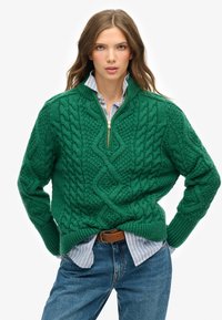 Suéter verde de punto trenzado con cuello de cremallera de un cuarto, patrones texturizados y puños acanalados, superpuesto a una camisa a rayas azul claro.
