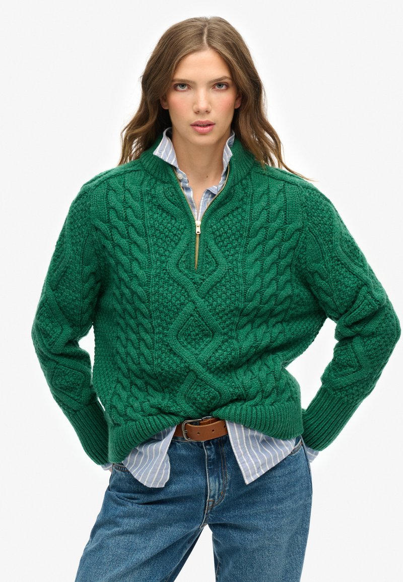 Suéter verde de punto trenzado con cuello de cremallera de un cuarto, patrones texturizados y puños acanalados, superpuesto a una camisa a rayas azul claro.