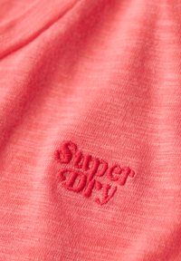 Korallpink stof med rødt broderet tekst "Super Dry" syet i en pæn, kursiv skrifttype på overfladen.