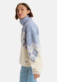 Pull en polaire bleu clair et crème, avec un col montant, des motifs géométriques et une étiquette de logo au bas. Texturé et confortable.