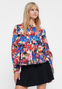 Woman wearing a multicolored floral ruffled blouse and a black pleated skirt, looking slightly to the right with hand resting on hip.