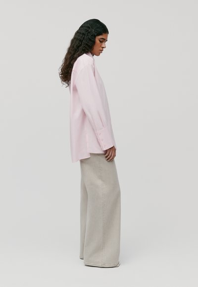 Camicia button-up rosa chiaro con vestibilità rilassata, maniche lunghe e spacchi laterali; abbinata a pantaloni ampi grigi chiari.