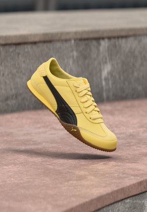 Baskets jaunes avec une bande noire et une semelle en gomme flottant au-dessus d'un trottoir rose et des marches en pierre grises en arrière-plan.