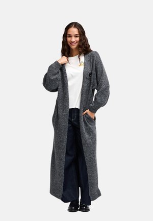 Jeune femme aux longs cheveux bouclés portant un long cardigan gris, un t-shirt blanc, un jean large foncé et des chaussures noires, souriant à la caméra.