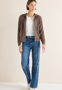 Brązowa bluza z kapturem zapinana na zamek z troczkiem, biała koszulka i niebieskie jeansy o prostych nogawkach. Białe sportowe buty dopełniają swobodną stylizację. Gładka faktura tkaniny.