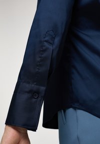 Camicia blu navy con una texture liscia e lucida, dotata di polsini con bottoni e un orlo leggermente curvato. Vestibilità sartoriale messa in evidenza nella silhouette.