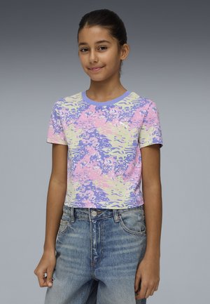 Meisje met donker haar naar achteren, draagt een t-shirt met een pastelpaars, roze en geel patroon en een high-waist blauwe spijkerbroek, staand tegen een grijze achtergrond.