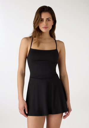 Donna che indossa un vestito nero senza maniche con spalline sottili e gonna svasata, in piedi davanti a uno sfondo bianco semplice.