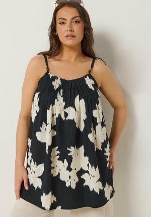 Vrouw draagt een zwarte mouwloze top met witte bloemenpatronen en dunne bandjes, staand tegen een effen lichtgekleurde muur.