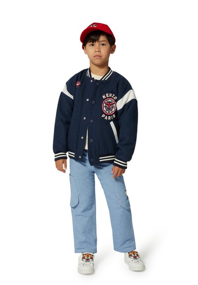 Navy varsity jacket with white accents and Kenzo logo, blue cargo pants, and white sneakers featuring a tiger design, finished with a red cap.