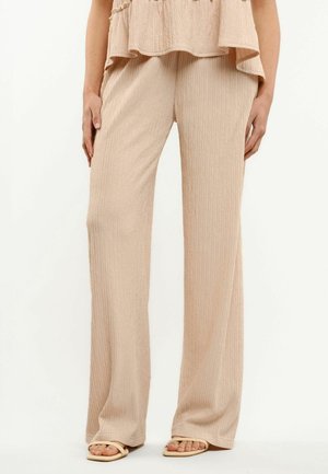 Žena v béžových žíhaných nohaviciach širokého strihu s nezlučujúcim sa topom a béžovými sandálmi s otvorenou špičkou, stojaca pred jednoduchým pozadím.