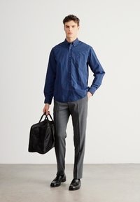 Cămașă bleumarin cu nasturi și finisaj texturat, pantaloni gri croiți, pantofi negri din piele și o geantă de voiaj neagră.