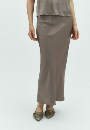 Femme portant une longue jupe taupe et des ballerines métalliques à bout pointu avec des détails de boucle, debout devant un fond clair uni.