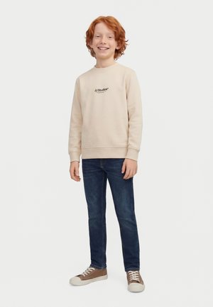 Rdečelasi fant se smeji, nosi bež pulover z napisom, temno modre kavbojke in rjave superge, stoji pred enobarvnim belim ozadjem.