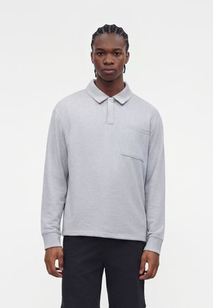 Homme aux cheveux tressés portant un polo gris clair à manches longues avec poche poitrine et pantalon noir, debout devant un fond uni.