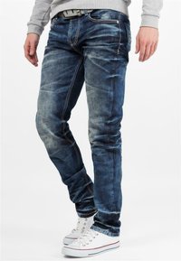 Jean en denim bleu skinny avec un léger effet usé, des coutures contrastantes et un design classique à cinq poches, associés à des baskets blanches.