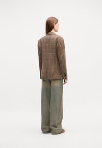Blazer marron à carreaux avec une coupe ajustée, associé à un pantalon large vert. Texture lisse ; pas de motifs visibles sur le pantalon.
