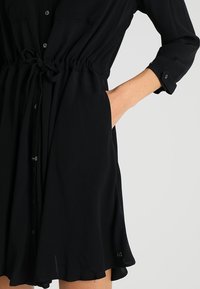 Person in einem schwarzen langärmeligen Kleid mit einer Kordelzug-Taille, einer eingetäuschten Tasche und hochgerollten Ärmeln vor einem einfarbigen Hintergrund.