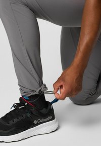 Pantalons de sport gris avec des bas ajustables, en train d'être resserrés par une main. Des chaussures de course noires avec des semelles blanches et un design texturé sont visibles.