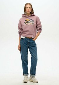 Pink hoodie with graphic print, loose-fitting blue jeans, and beige sneakers. Hoodie features a kangaroo pocket and ribbed cuffs.
