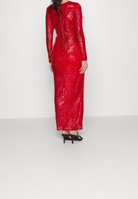 Rotes Pailletten-Kleid mit langen Ärmeln, bodenlangem Design und einem Reißverschluss auf der Rückseite. Getragen mit schwarzen High Heels.