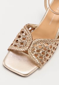 Sandale avec bride en cuir tissé doré et bout métallique carré. Présente des accents tressés et une surface lisse et brillante au niveau du talon et de la semelle.