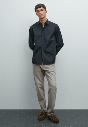 Junger Mann, der mit den Händen hinter dem Rücken steht und ein schwarzes Hemd mit Knöpfen, hellbeige Hosen und braune Wildlederschuhe trägt.