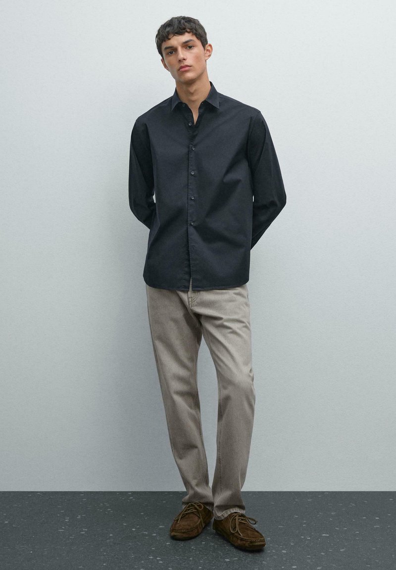 Giovane in piedi con le mani dietro la schiena, indossa una camicia nera con bottoni, pantaloni beige chiaro e scarpe in suede marroni.