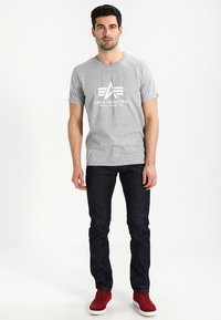 Gray cotton t-shirt with a crew neck, featuring a white logo print. Worn with dark blue jeans and red sneakers. Smooth texture and casual fit.