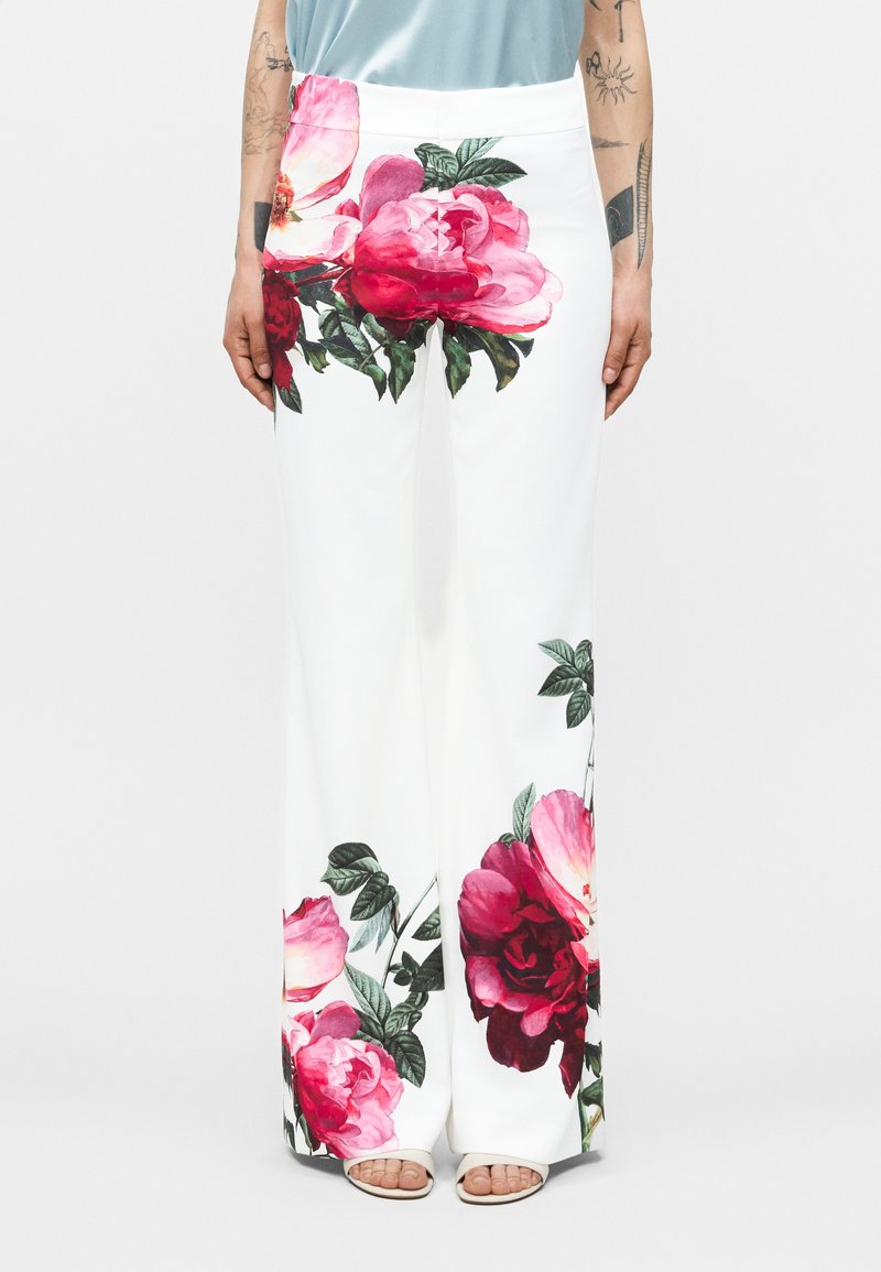 Pantalones blancos de pierna ancha con grandes estampados florales en rosa y rojo en las caderas y la parte baja de las piernas, combinados con sandalias de punta abierta y una blusa azul claro.