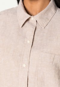 Lys beige linned skjorte med krave, frontknaplukning og en enkelt brystlomme. Stoffet er tekstureret med et subtilt vævemønster.