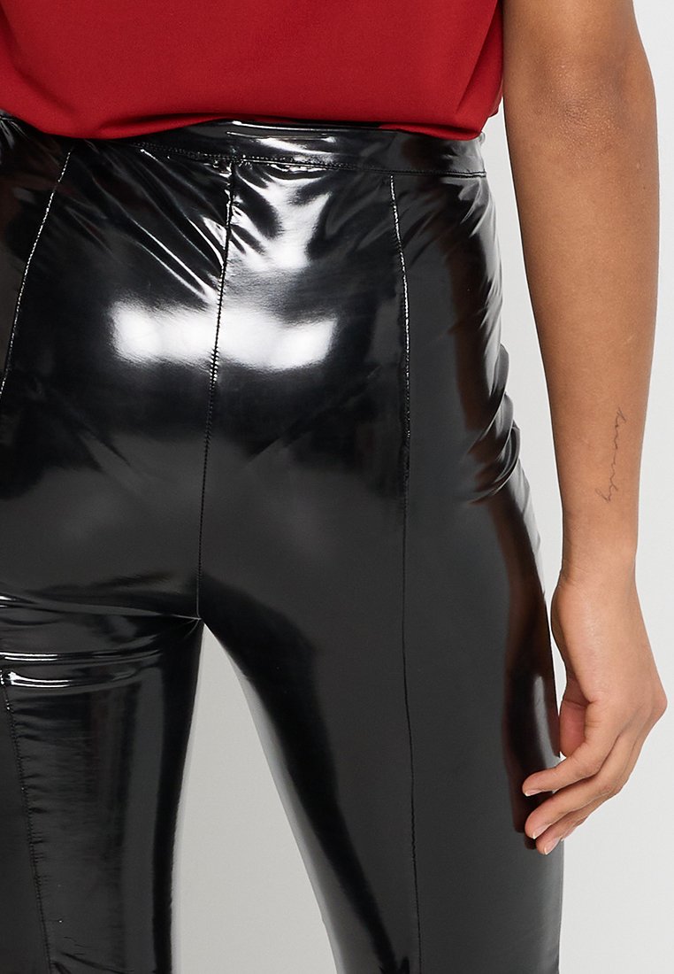 Hoch-taillierte schwarze glänzende Leggings aus schimmerndem Material, mit glatter Textur und Nahtdetail vorne, kombiniert mit einem bordeauxfarbenen Oberteil.