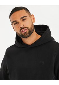 Zwarte hoodie van zacht fleece materiaal. Heeft een voorzak en een subtiele logo op de borst. Eenvoudig, ontspannen ontwerp.