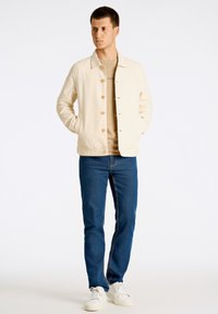 Ljusbeige bomullsjacka med knappstängning, sidofickor och en avslappnad passform, tillsammans med blå jeans och vita sneakers.