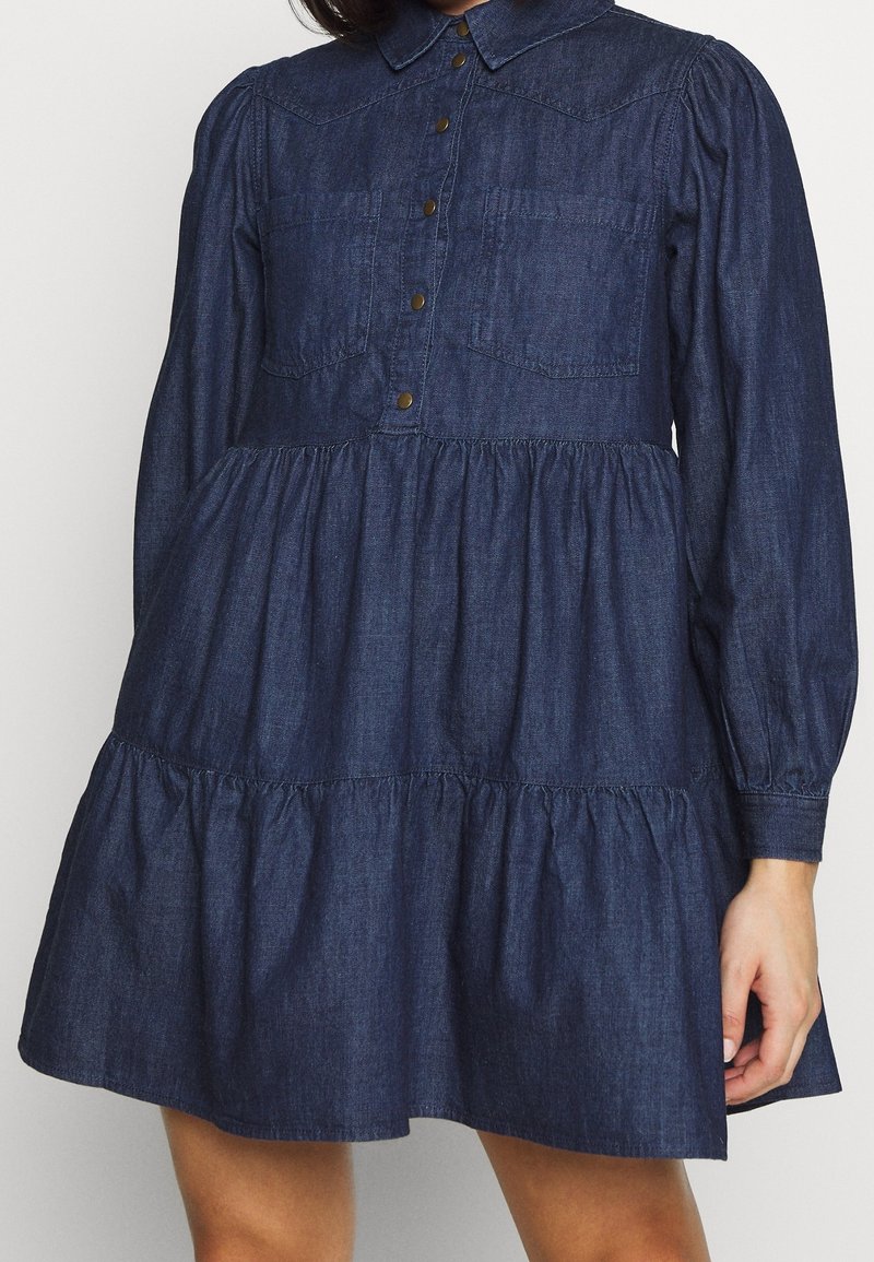 Denim-mekko, jossa on napitettu etuosa, kaulus, pitkät puffihihat, kaksi rintataskua ja kerroksellinen, liehuva helma tummansinisenä.